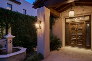 Wooden front door with ornate panels under a covered entry, illuminated by hanging lanterns, ivy-clad walls, and a tiled stone courtyard at dusk.